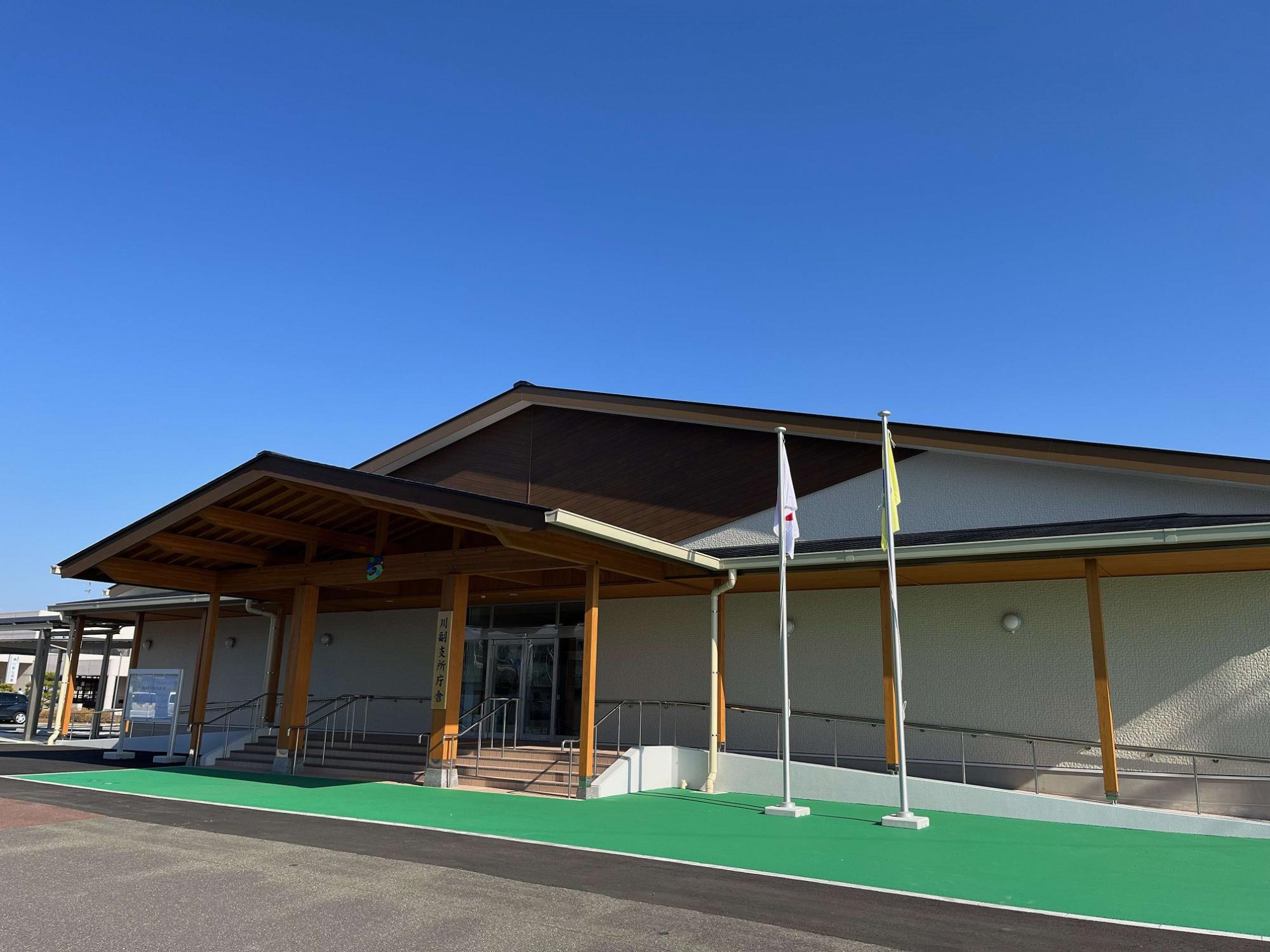 木材を活かした大きな屋根と、スロープを備えたバリアフリー設計が特徴的な平屋建ての建物で手前には緑色の歩行路と二本の旗ポールが設置された川副支所庁舎の外観写真