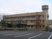 アスファルトが敷かれた駐車場の奥に建つ、ベージュ色をした三階建ての建物と、その右端にそびえ立つ円筒形の展望塔のような構造物を捉えた東与賀支所の外観写真