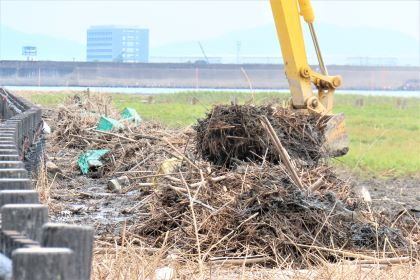 黄色い重機のアームが干潟に積まれた大量の漂着ゴミや枯れ草をバケットで掴み上げており、背景には遠くのビルや堤防が広がる中で清掃作業が行われている様子の令和7年8月28日時点の写真
