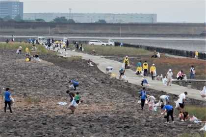 海岸沿いの干潟のような岩場で、青や黄色のシャツを着た大勢のボランティアの人々が、白いゴミ袋を手に持ちながら一つひとつ丁寧にゴミを拾い集めて清掃活動に励んでいる様子を捉えた、令和7年9月28日時点の写真