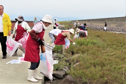 ボランティアの人々が、海岸の舗装路周辺に散乱した大量の流木や細かな枝を丁寧に拾い集め、指定の大きなゴミ袋に入れて環境保全活動に協力している様子の令和7年9月28日時点の写真