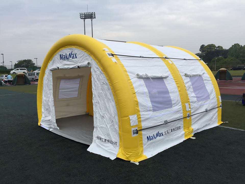 屋外のグラウンドに設営された、黄色い空気注入式のフレームと白い膜材で構成された「MakMax 太陽工業株式会社」のロゴ入り大型エアーテントを、斜め前方からその構造が分かるように捉えた写真
