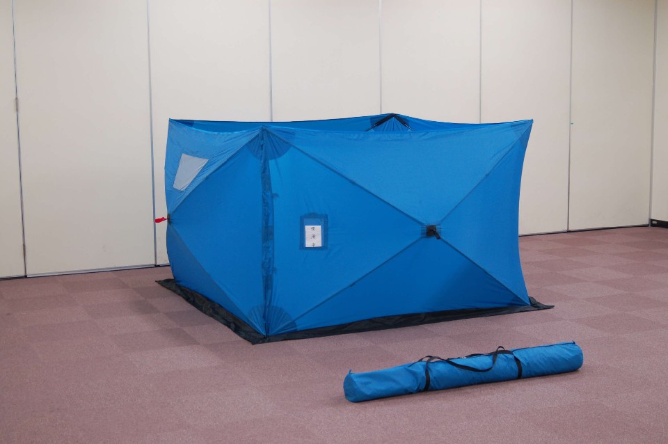 屋内のカーペット敷きのスペースに設置された、プライバシー保護や避難所での利用を目的とした青いナイロン製のワンタッチ式パーティションと、その手前に置かれた専用の細長いキャリーバッグを斜めから捉えた写真