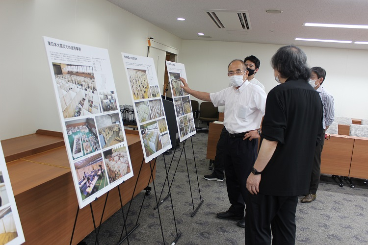 「東日本大震災での活用事例」などの文字と活動写真が掲載された複数の大きな展示パネルを前に、マスクを着用した男性が指をさして説明を行い、周囲の出席者がその内容を真剣な表情で聞き入っている説明会の様子を捉えた写真