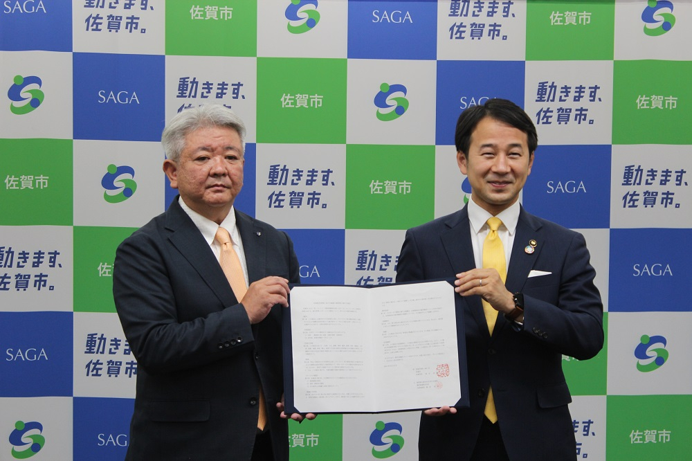 「動きます、佐賀市。」のキャッチコピーと市章が並ぶバックパネルの前で、揃いの黄色いネクタイを締めた二人の男性が、署名・捺印された協定書を正面に向けて掲げ、連携強化や合意の成立を報告するために撮影された写真