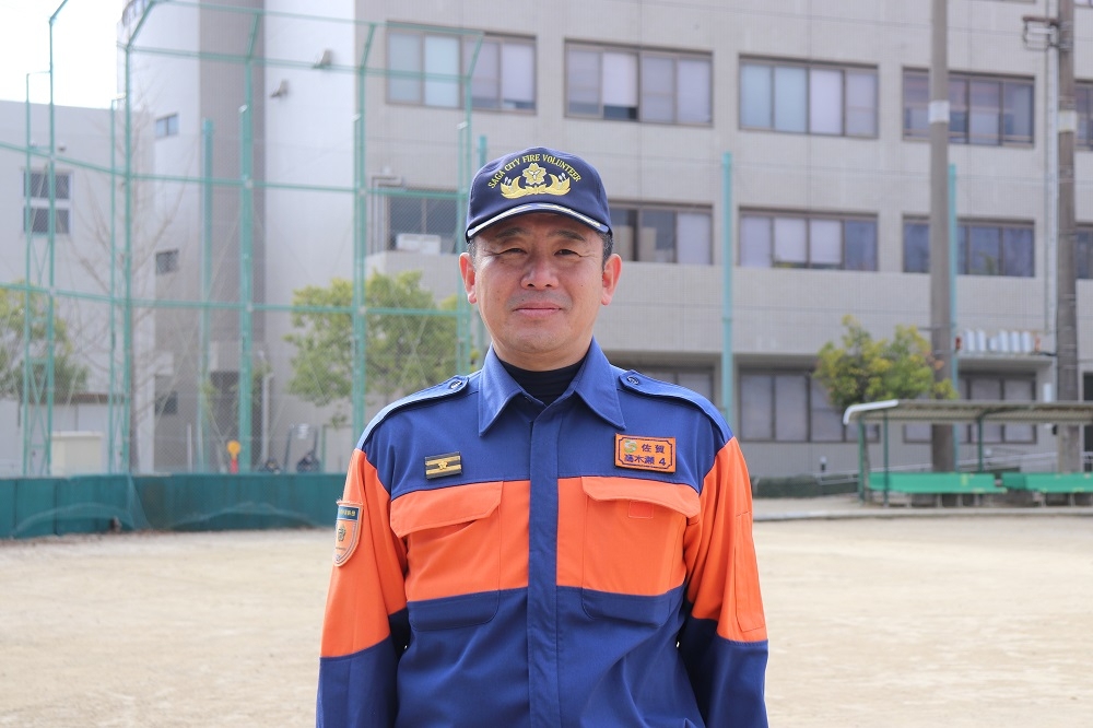 建物の前にある砂の訓練場で、紺とオレンジの配色が特徴的な消防団の制服と帽子を着用した山崎修司さんが穏やかな表情で正面を見据えて立っており、その胸元には佐賀市などの所属を示すワッペンや階級章が付けられている写真