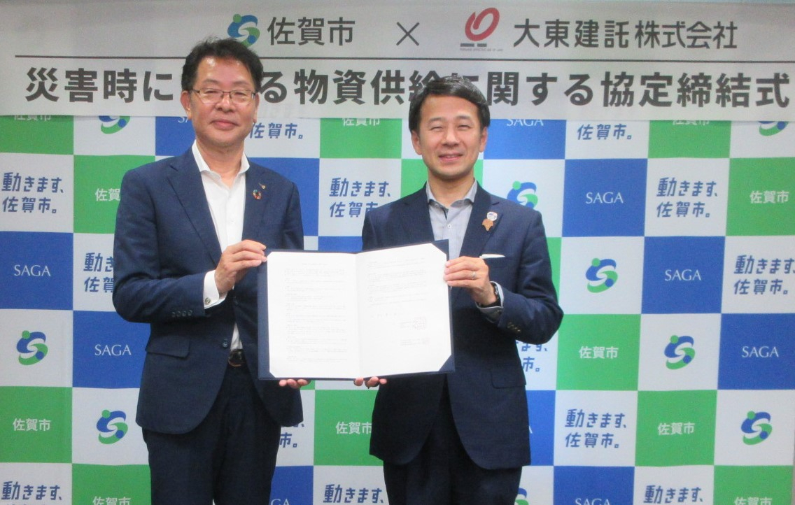 佐賀市と大東建託株式会社による「災害時における物資供給に関する協定締結式」において、スーツ姿の男性二人が協定書を手に持ち、市章やロゴが並ぶバックパネルの前で笑顔で並び立つ、協力関係の構築を記録した記念写真