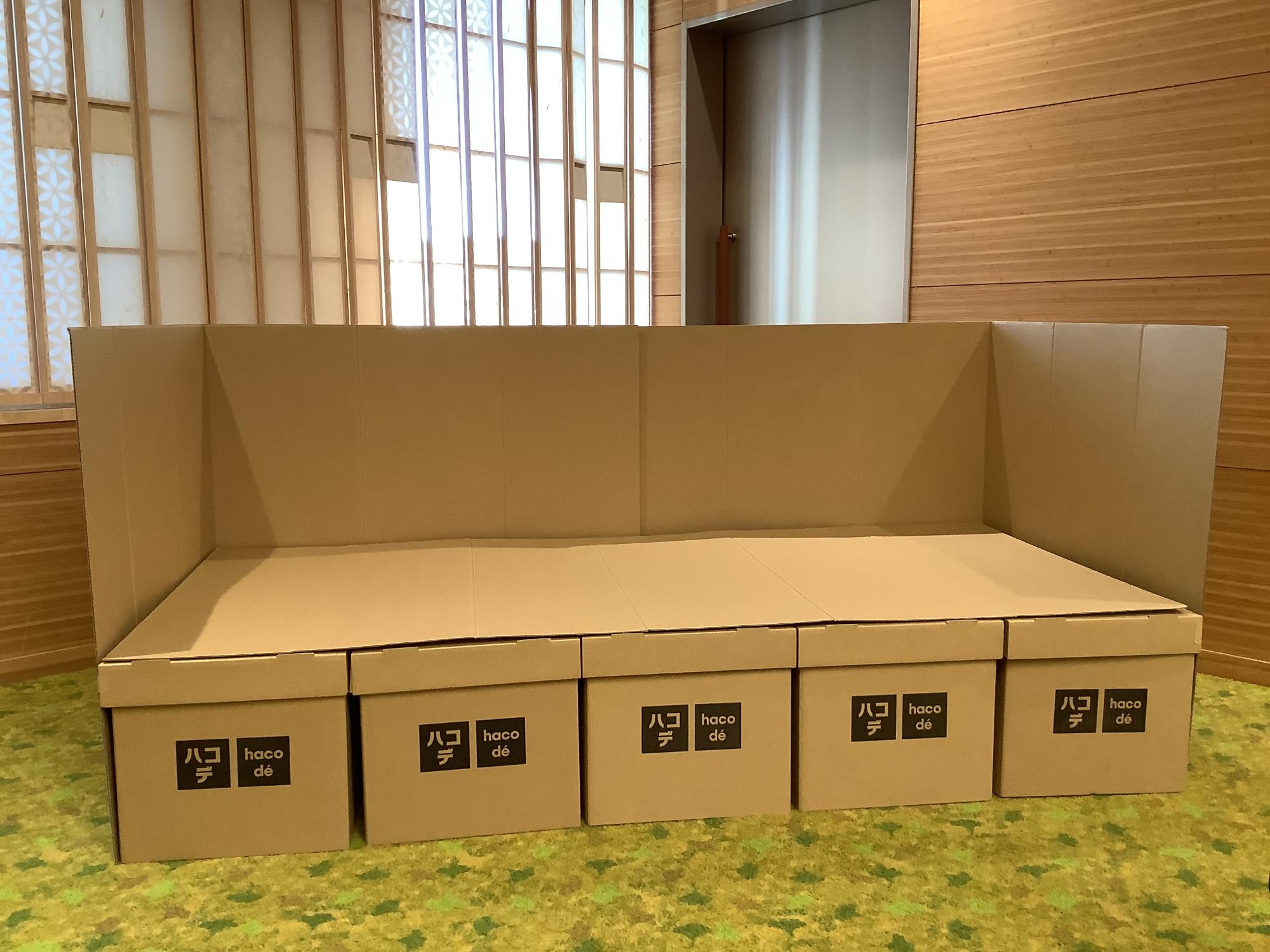前面に「ハコデ haco d&eacute;」のロゴが並ぶ5つの段ボール箱を土台にし、三方を高い仕切り壁で囲った段ボール製の簡易ベッド「ハコデベッド」が設置されている様子を写した写真