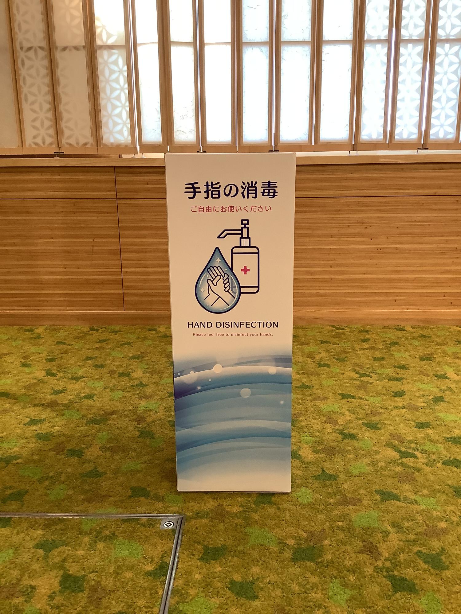 上部に「手指の消毒」という文字、中央に消毒液のポンプと手のイラスト、下部に青い水面のグラフィックが描かれた、ダンボール製消毒液スタンドを写した写真