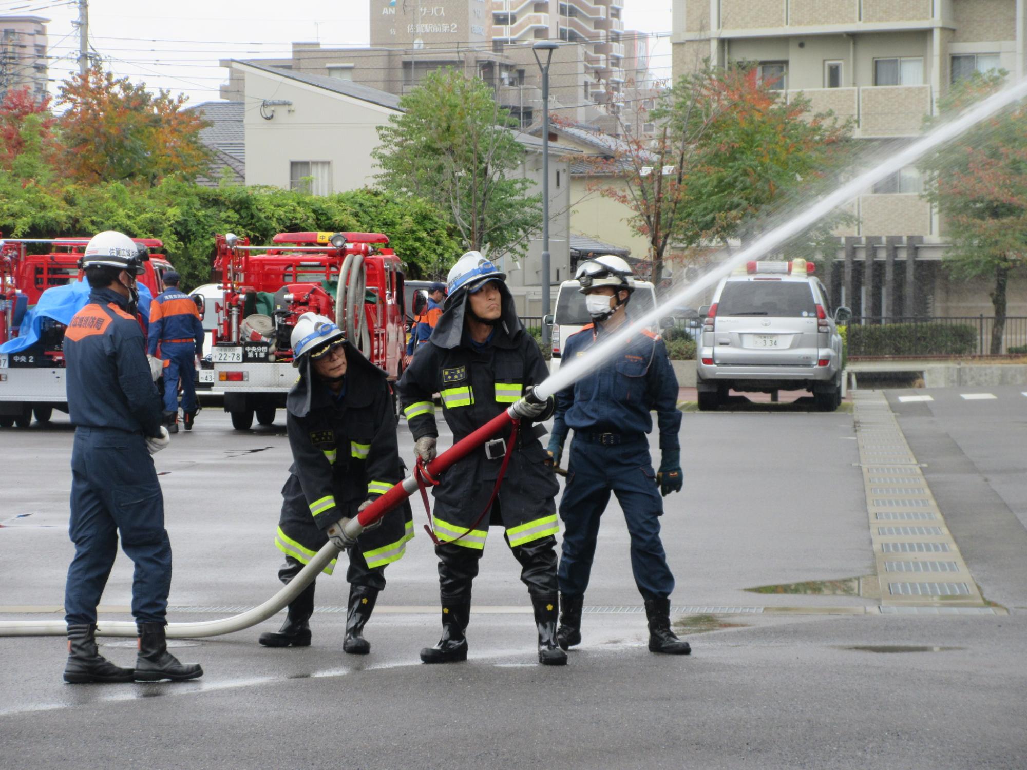 屋外の訓練場で紺色の防火服に身を包んだ3人の消防士が連携して太いホースを支えながら前方へ勢いよく放水しており、その後方では赤い消防車や別の隊員たちが活動を見守っている様子の写真