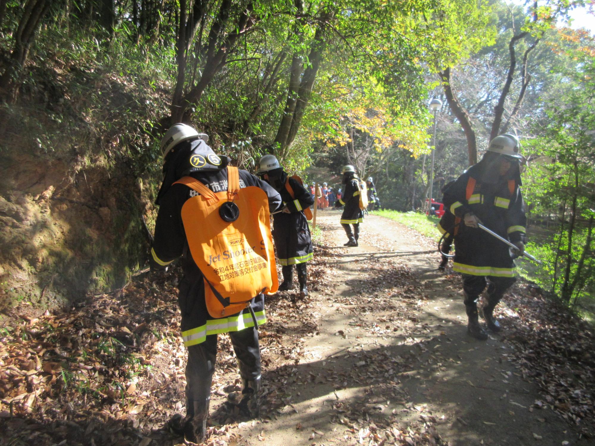 木々が茂る山道で防火服を着用した複数の消防団員が活動しており、手前の隊員がオレンジ色の消火用水の嚢を背負い、周囲の地面や斜面に向かって消火や点検作業を行う様子の写真