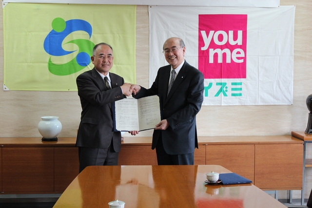 左側に黄緑色の市の紋章旗、右側に「you me イズミ」のロゴ旗が掲げられた室内で、スーツ姿の二人の男性が笑顔で握手を交わしながら見開いた協定書を共同で持ち、地域連携や事業協力の締結を記念して撮影された写真