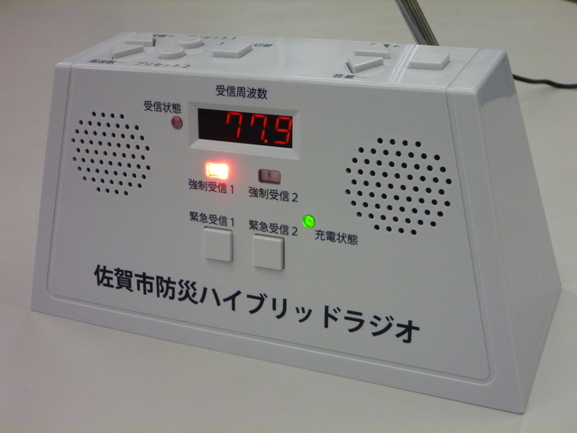 「佐賀市防災ハイブリッドラジオ」と印字された、前面にデジタル周波数表示や緊急受信ボタン、スピーカーを備えた白い卓上型ラジオが、電源ランプを点灯させて机の上に置かれている様子を斜め前から捉えた写真