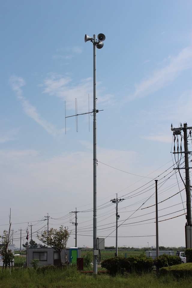 青空を背景に、先端に3方向を向いたスピーカーと通信用アンテナが設置された、災害時の避難勧告や緊急情報を住民に知らせるための背の高い防災行政無線の鉄塔を、周囲の電柱や畑と共に捉えた写真